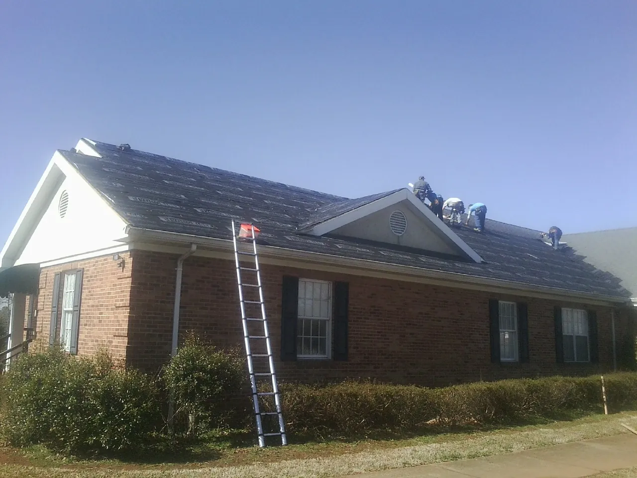 Crew tearing shingles off a brick ranch roof with a ladder set — Emergency Roof Repair work by our crew in Martensdale