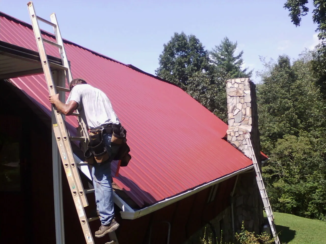 Roofer on a ladder beside a red metal roof and stone chimney — Soffit & Fascia Repair by Roofing Brothers in Martensdale